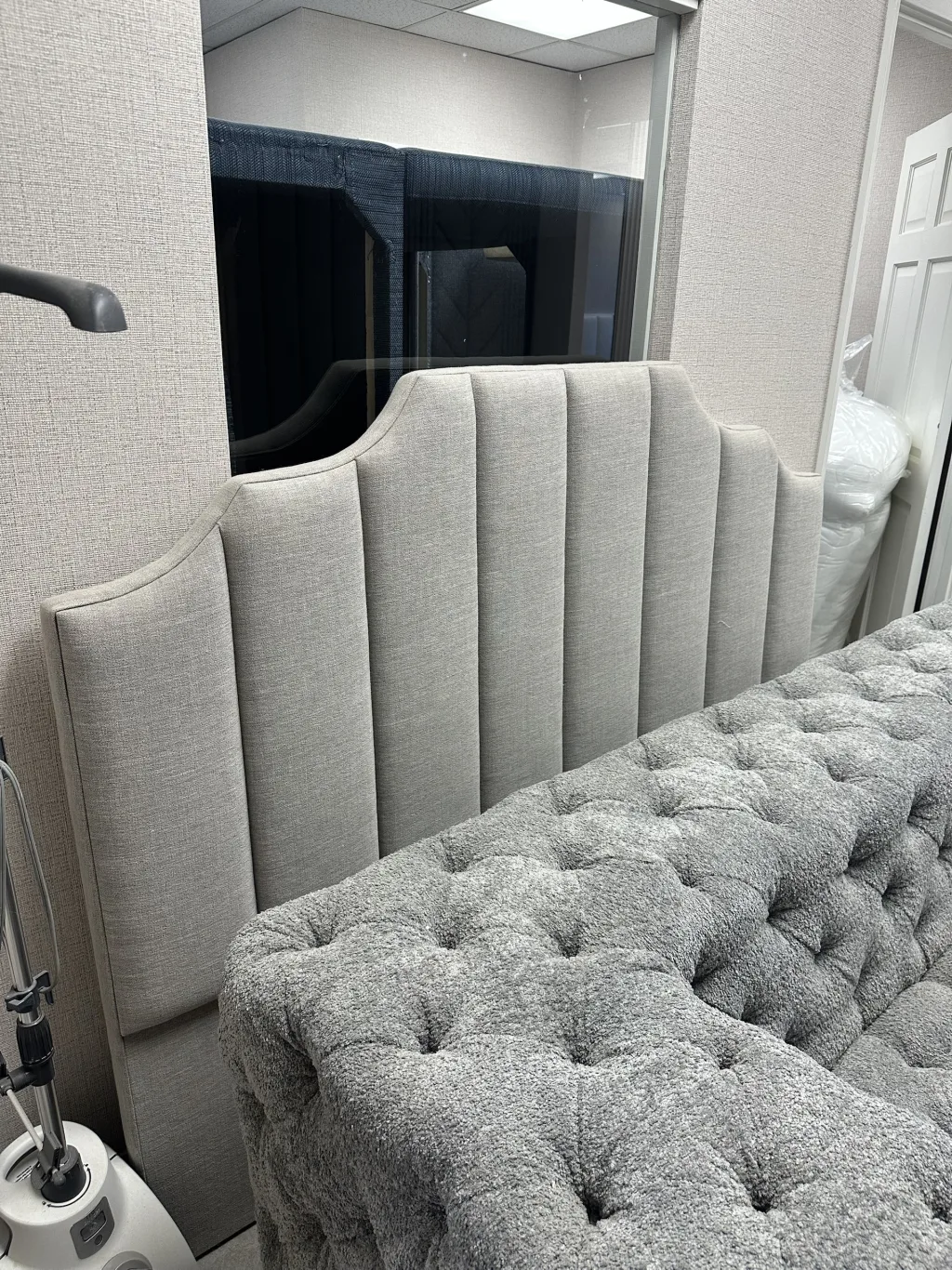 A pale grey channel-tufted headboard with a shaped stepped top, standing against a linen-panelled wall in the Elite showroom