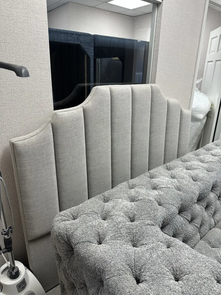 A pale grey channel-tufted headboard with a shaped stepped top, standing against a linen-panelled wall in the Elite showroom
