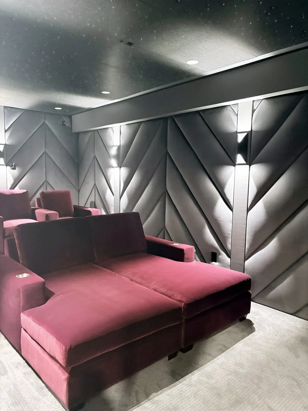 A deep red velvet chaise and a back row of recliners against a wall of grey chevron panelling, under a star-lit ceiling