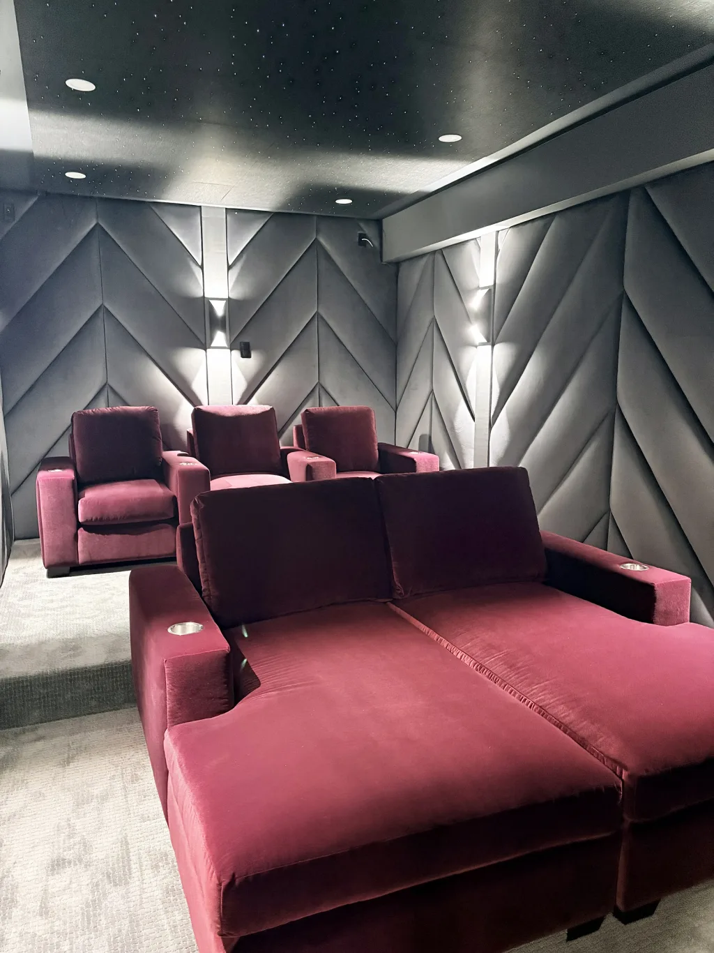 The same theatre room from the back corner, both rows of red velvet seating between two panelled walls lit by narrow sconces