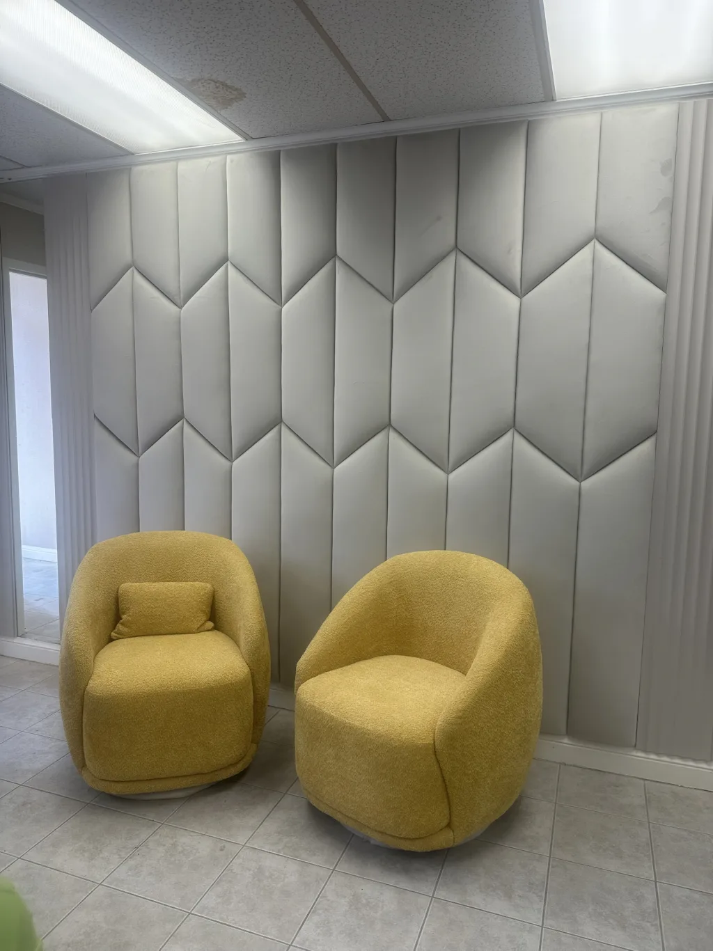 A wall of pale grey chevron-tufted panelling running the full height of the room, with two mustard bouclé swivel chairs in front of it