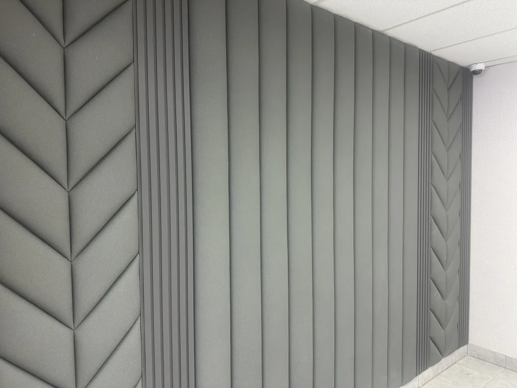 A wall of sage upholstered panelling running floor to ceiling, chevron sections meeting plain vertical channels and a narrow slatted band
