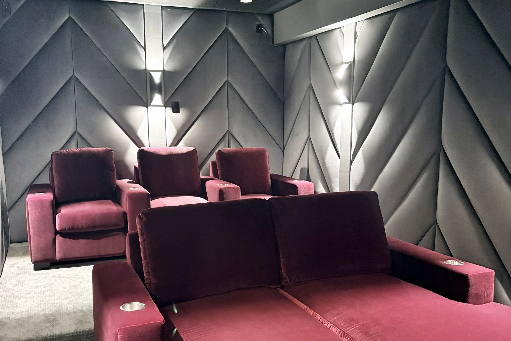 A home theatre Elite built, seen from the back corner: a deep red velvet chaise in front of a row of matching recliners, against a wall of grey chevron acoustic panelling lit by narrow sconces